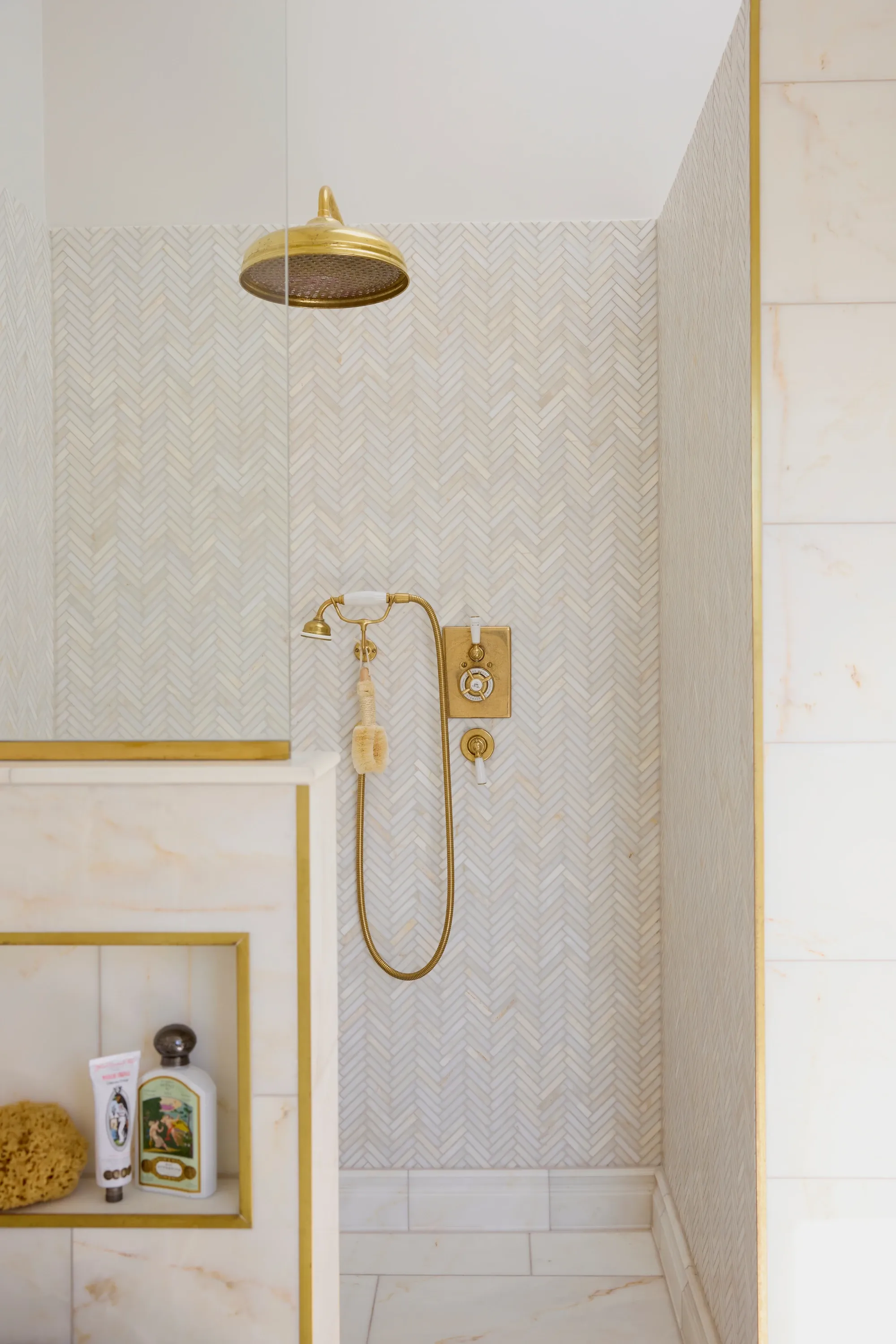 A brass rainfall shower head and handset on a wall of pale herringbone mosaic tiles, beside a marble half wall with a brass trimmed niche.