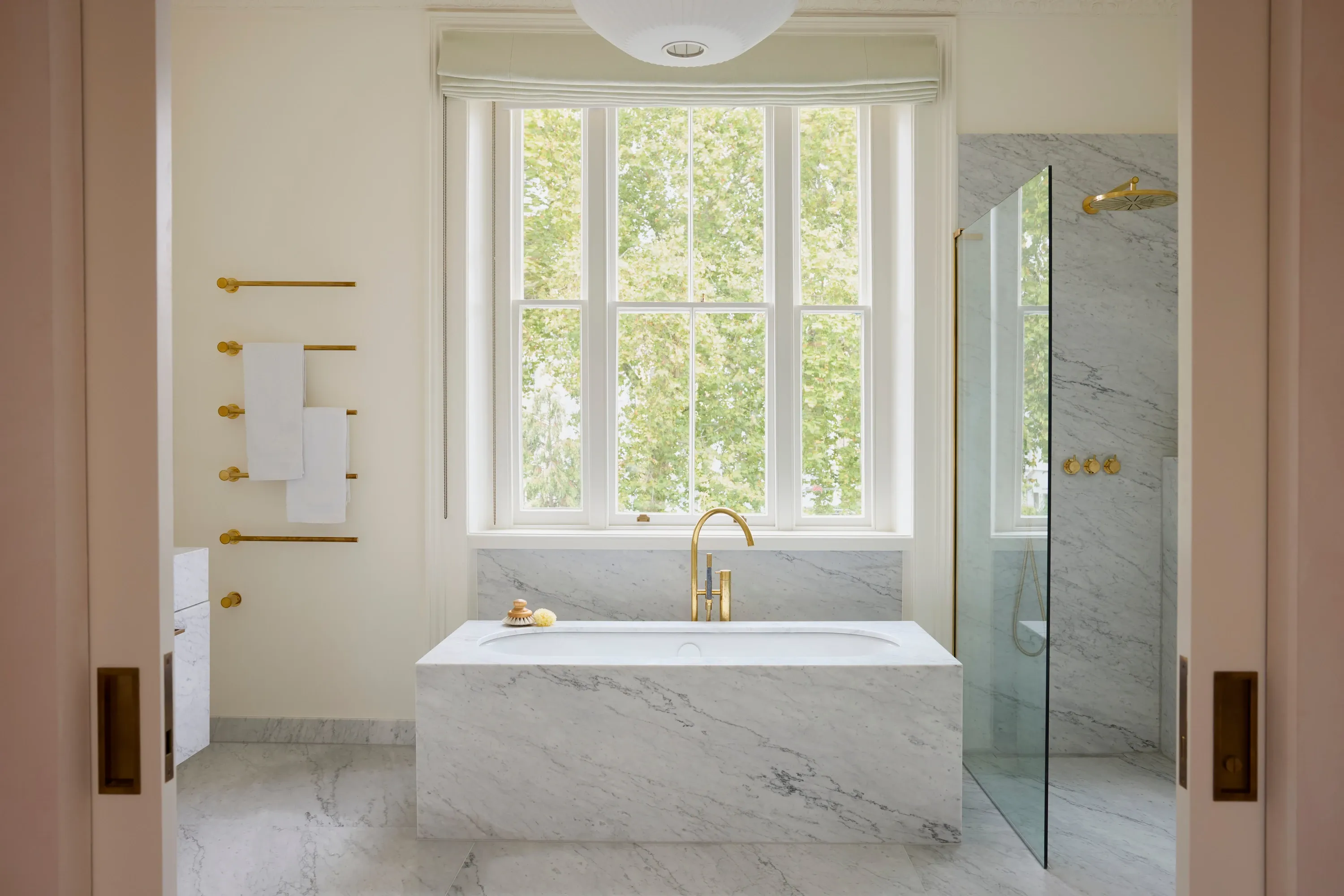 A marble clad bath sits beneath a tall bay window with a brass floor tap, beside a marble walk-in shower with a brass rainfall head.