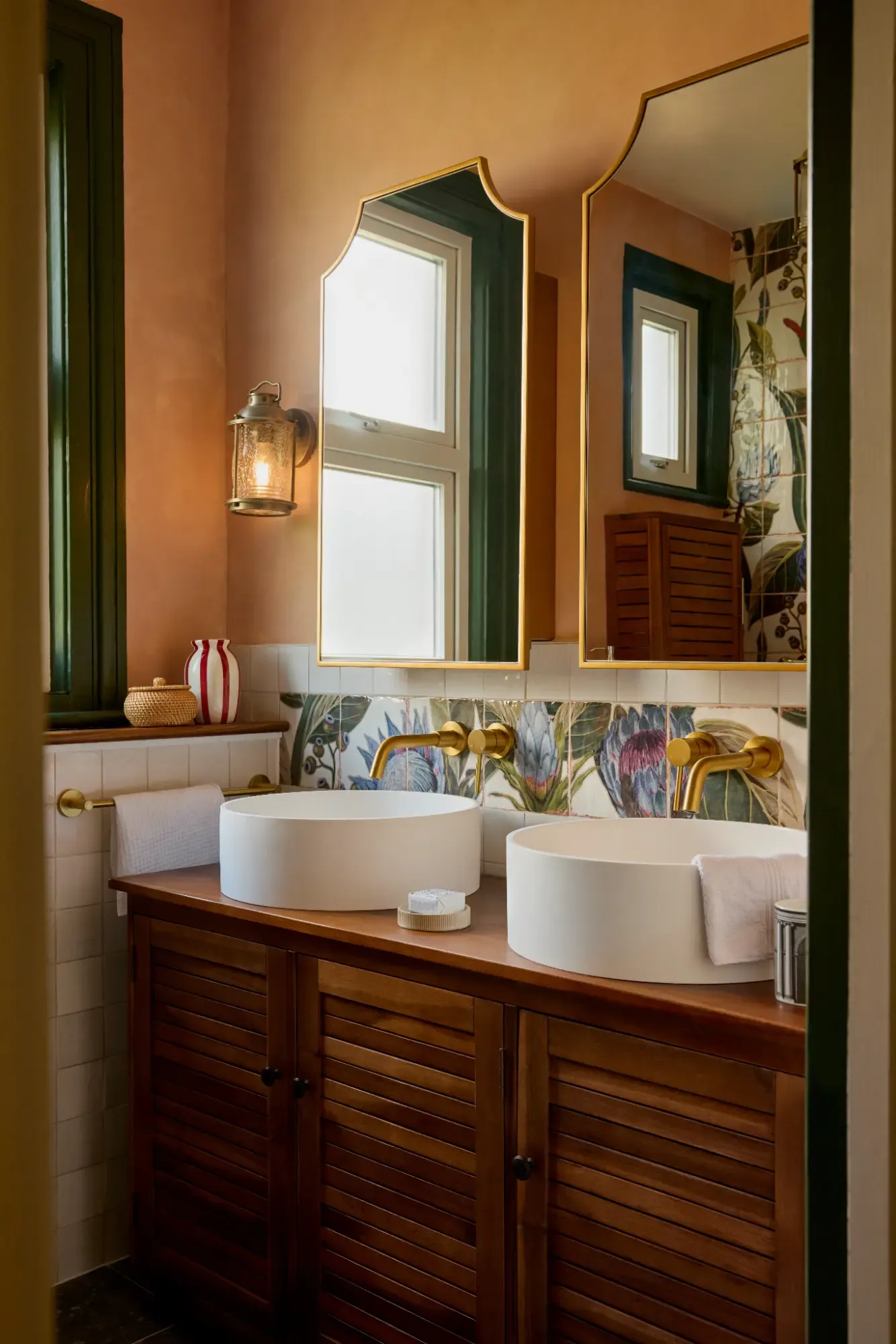 Two round white basins on a dark wood louvred vanity with brass wall taps, below a band of hand painted botanical tiles and terracotta walls.